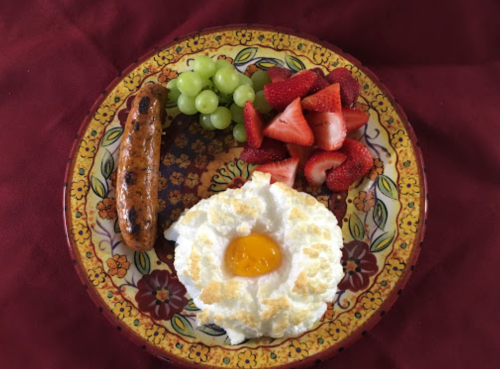 A colorful plate with a sunny-side-up egg, sausage, green grapes, and sliced strawberries on a decorative background.