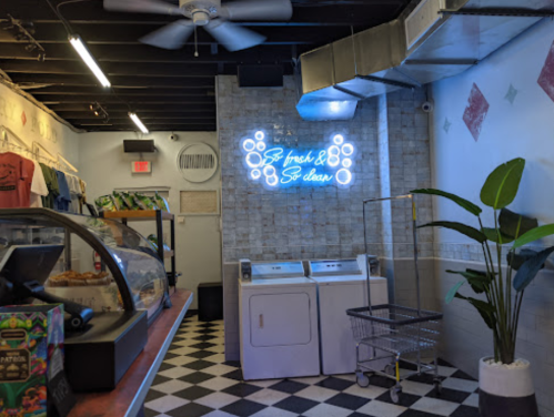 A cozy interior of a cafe featuring a neon sign, checkered floor, and a laundry area with a plant in the corner.