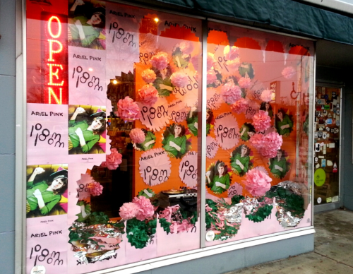A shop window decorated with colorful posters of Ariel Pink and large paper flowers, with an "OPEN" sign illuminated.
