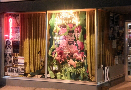 A brightly lit storefront window displays colorful decorations and posters for the Flamingo Hotel.