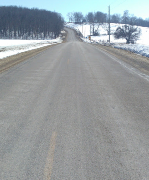 A snow-covered rural road stretches into the distance, flanked by trees and fields under a clear blue sky.