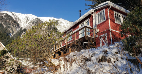 A red house on a snowy hillside, surrounded by trees, with mountains in the background under a clear blue sky.
