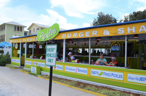 A colorful restaurant named "Pickles" with outdoor seating, serving burgers and shakes, surrounded by greenery.