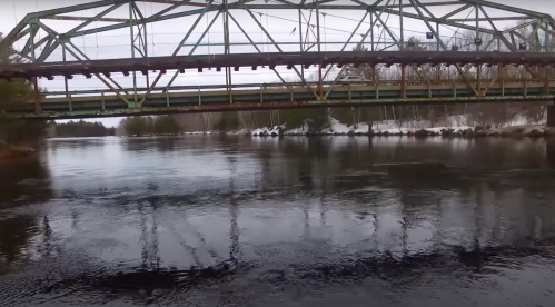 A weathered metal bridge spans a calm river, reflecting its structure in the water, surrounded by snow-covered banks.