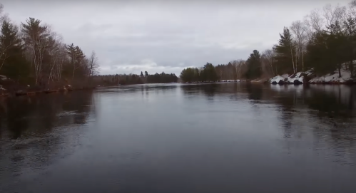 A calm river scene surrounded by trees, with a cloudy sky and snow on the banks.