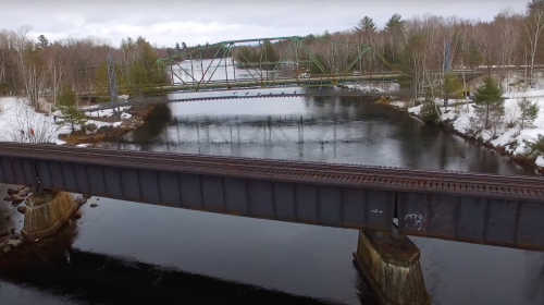 Aerial view of two bridges over a river, surrounded by trees and snow, with a cloudy sky above.