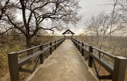 A wooden walkway leads to a gazebo, surrounded by bare trees and a cloudy sky.
