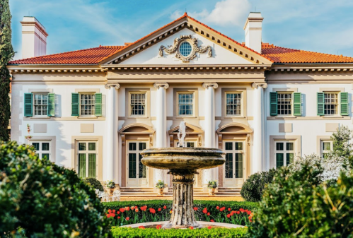 A grand mansion with a fountain in front, surrounded by manicured gardens and vibrant flowers under a clear blue sky.