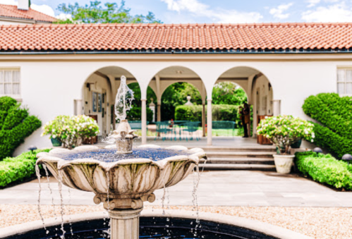 A decorative fountain in the foreground with a lush garden and a house featuring arched openings in the background.