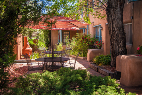 A cozy outdoor patio with a table, chairs, and a large umbrella surrounded by greenery and colorful flowers.