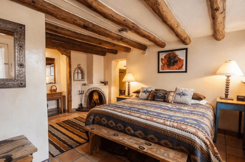 Cozy bedroom with a wooden beam ceiling, a patterned bedspread, and a fireplace, featuring warm lighting and rustic decor.