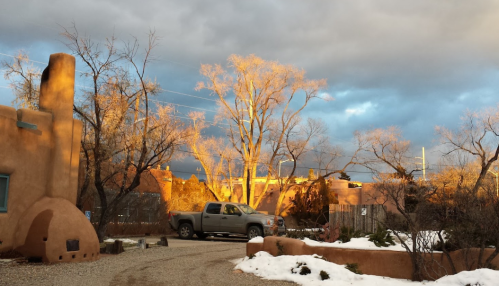 A dirt road leads past adobe buildings and bare trees, illuminated by warm sunset light against a cloudy sky.