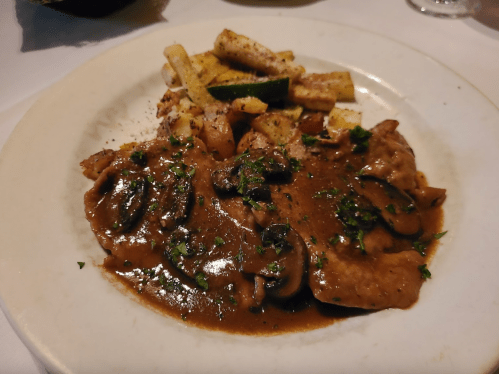 A plate of sliced meat in mushroom sauce, garnished with herbs, served with roasted vegetables and fries.