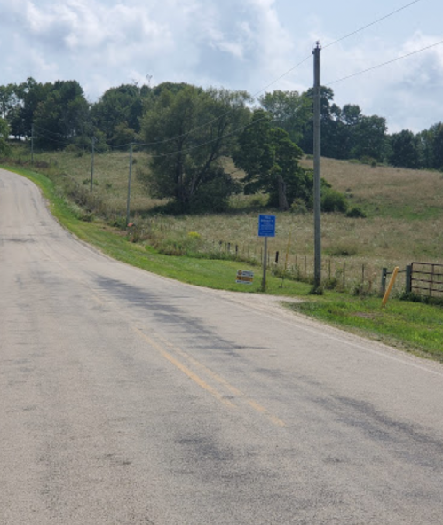 A rural road curves upward, flanked by grassy fields and trees, with a sign visible on the side.