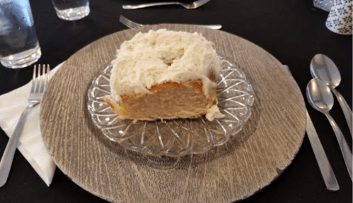 A slice of cake with white frosting on a decorative glass plate, set on a textured gray table.