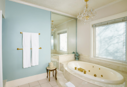 A bright bathroom featuring a large tub, a mirror, and white towels hanging on a gold rack against a blue wall.