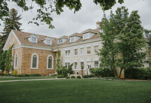 A large, elegant brick and stucco building surrounded by lush greenery and trees under a cloudy sky.