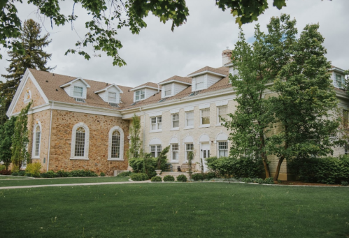 A large, elegant brick and stucco building surrounded by lush greenery and trees under a cloudy sky.