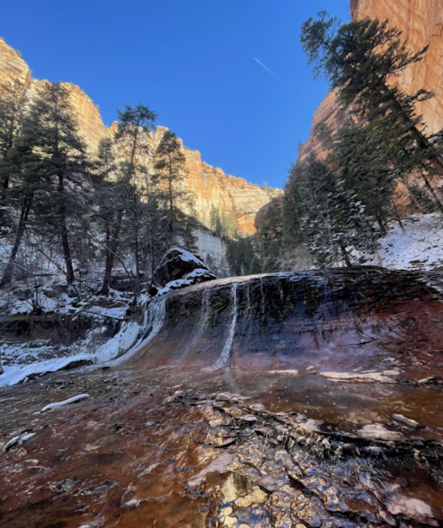 A serene waterfall cascades over a rocky ledge, surrounded by snow-dusted trees and towering cliffs under a clear blue sky.
