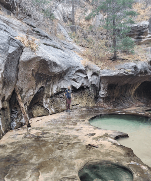 A person stands on a rocky surface near a natural pool, surrounded by steep rock formations and sparse trees.