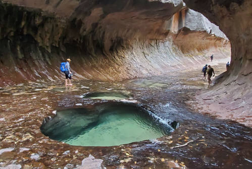 Hikers explore a narrow canyon with smooth rock walls and clear pools of water along the riverbed.