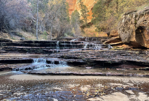 A serene landscape featuring cascading waterfalls over rocky terrain, surrounded by trees and autumn foliage.