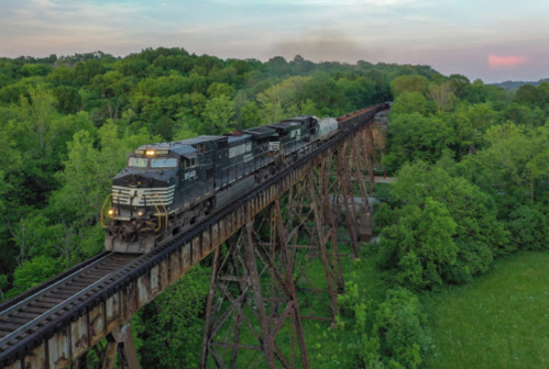 A black train crosses a tall bridge surrounded by lush green trees at sunset.