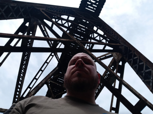 A person stands beneath a rusted metal structure, looking up at the intricate framework against a cloudy sky.