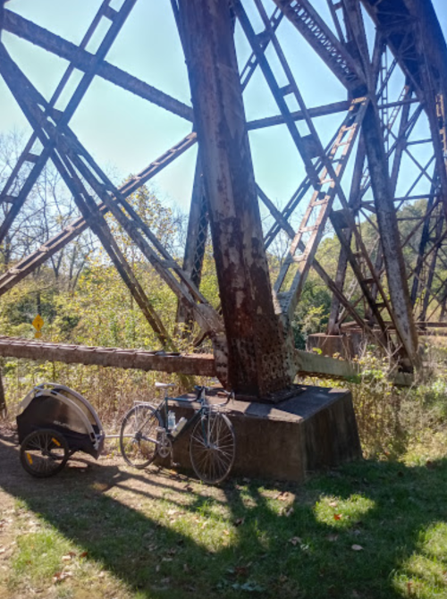 A bicycle and a bike trailer are parked under a large, rusted metal structure surrounded by greenery.