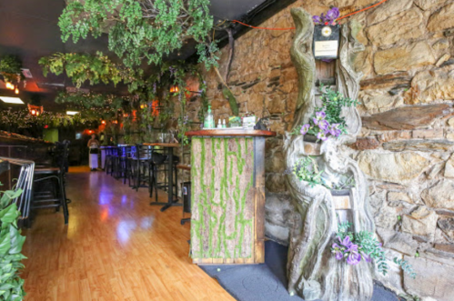 A cozy restaurant interior with stone walls, wooden decor, and lush greenery, featuring a bar and seating area.