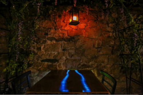 A cozy stone wall with a glowing lantern and a table reflecting blue light, surrounded by greenery.