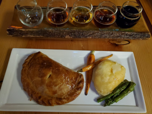 A plate with a golden pastry, mashed potatoes, carrots, asparagus, and a flight of four small glasses of different drinks.