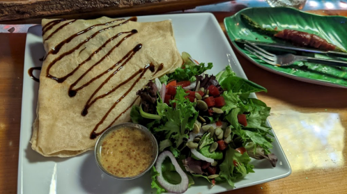 A plate with a crepe drizzled with sauce, accompanied by a fresh salad and a small bowl of mustard.