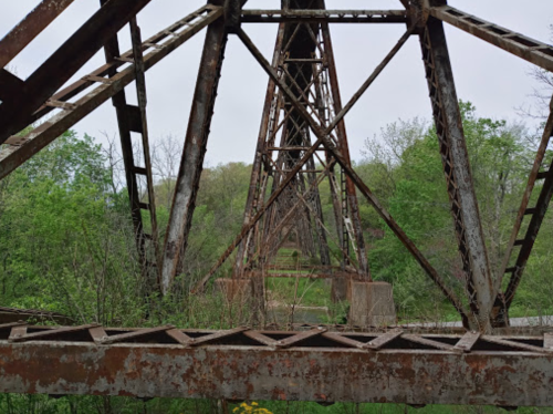 Rusty metal framework of an old bridge, surrounded by lush green trees and foliage.