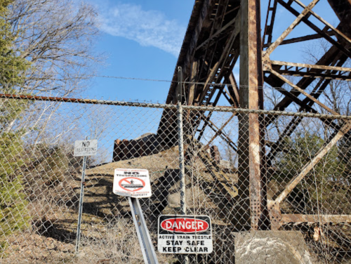 A rusted train trestle viewed through a chain-link fence with warning signs about train activity and safety.