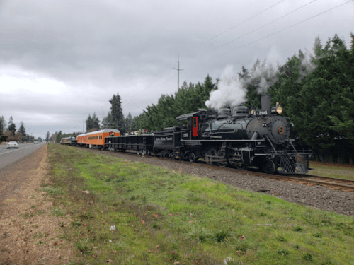 A vintage steam train with a black locomotive and orange passenger cars travels along a track beside a road.