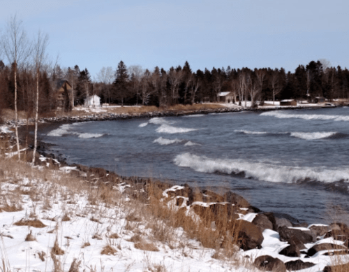 A winter shoreline with snow-covered rocks, gentle waves, and trees in the background under a clear blue sky.