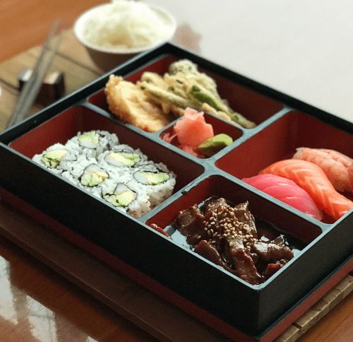 A bento box with sushi, tempura, rice, and teriyaki beef, served with chopsticks on a wooden table.