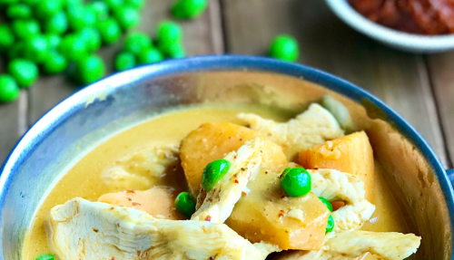 A bowl of chicken curry with green peas and chunks of potato, surrounded by green peas and a small bowl of sauce.