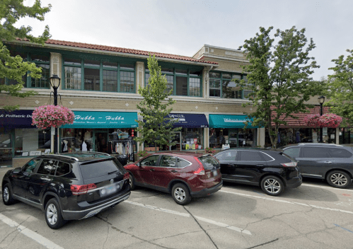 A street view of shops with hanging flower baskets and parked cars in front of a two-story building.