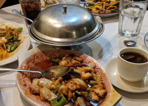 A table set with a plate of chicken dish, a covered bowl, a cup of tea, and other dishes in the background.