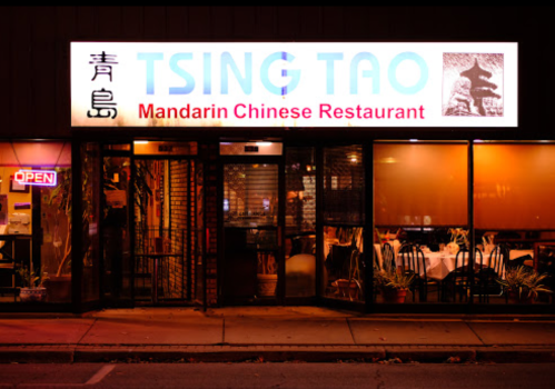 Exterior of Tsing Tao Mandarin Chinese Restaurant, featuring a lit sign and open door, with tables visible inside.