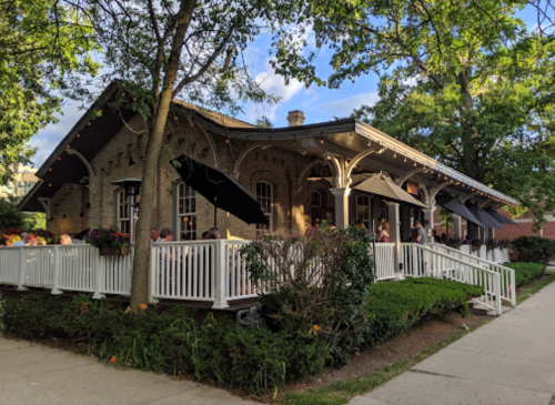 A charming brick restaurant with outdoor seating, surrounded by greenery and trees on a sunny day.