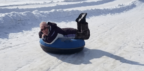 A person in winter clothing lies on a blue snow tube, smiling as they slide down a snowy slope.