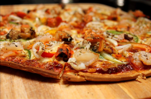 A close-up of a seafood pizza topped with shrimp, vegetables, and herbs on a wooden cutting board.