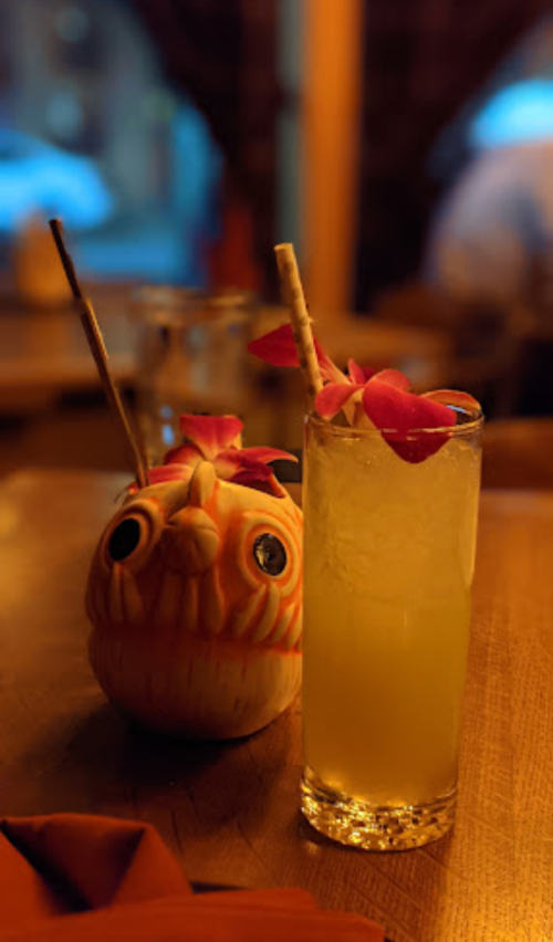 A colorful drink garnished with flowers next to a carved fruit figure resembling an owl, set in a warm-lit environment.