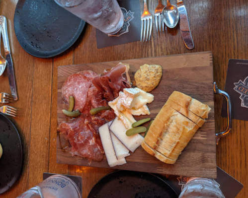A wooden platter with assorted meats, cheese, pickles, and sliced bread, set on a table with glasses and plates.