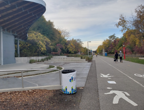 A paved path alongside a park with trees, a trash bin, and people walking in the distance under a cloudy sky.