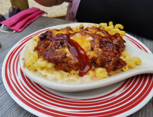 A dish of macaroni and cheese topped with barbecue chicken and drizzled with sauce, served in a white bowl on a red plate.