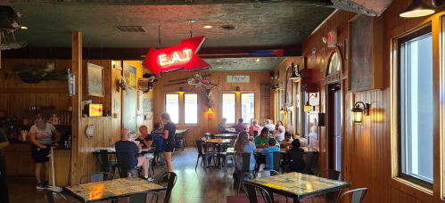 Interior of a rustic restaurant with wooden walls, tables, and a neon "EAT" sign, filled with diners and staff.